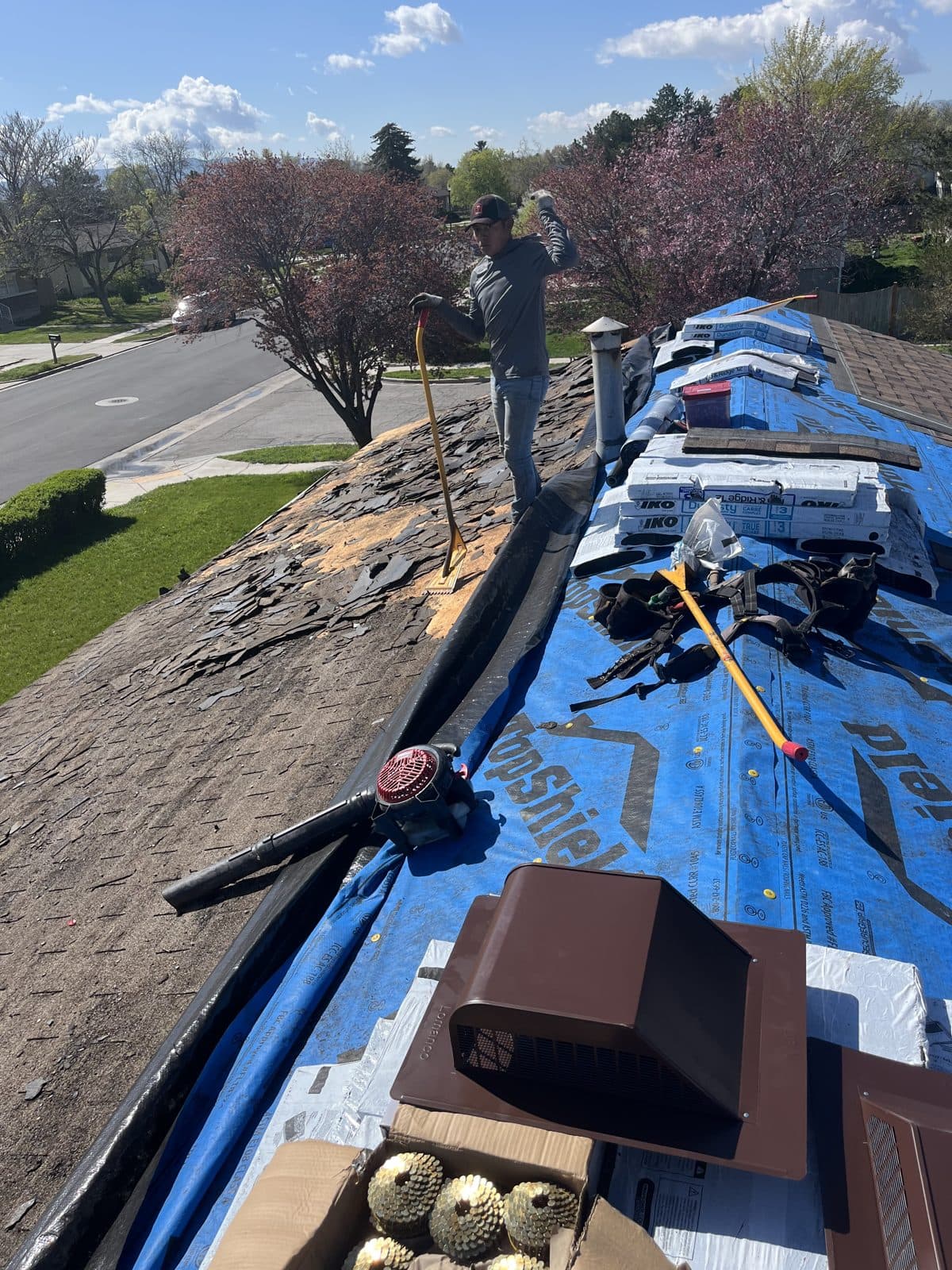 Tear-off on one side of a roof and synthetic underlayment with shingle bundles on the other, with a worker holding a shovel.